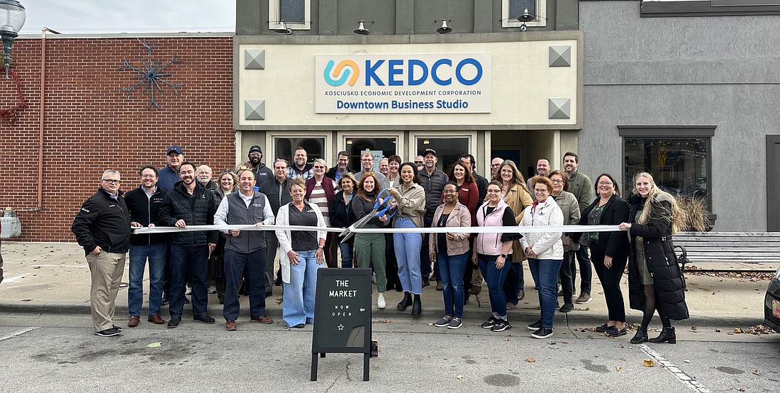 Market Associates Laura Sanders and Melanie Woodruff (center, with scissors) for The Market at KEDCO Downtown Business Studio cut the ribbon during the ribbon-cutting ceremony with the Kosciusko Chamber of Commerce. Also pictured are Chamber President and CEO Rob Parker (first row, left), Chamber ambassadors, Main Street Warsaw members, city and county officials and KEDCO CEO Alan Tio (first row, fourth from left). Photo by David Slone, Times-Union
